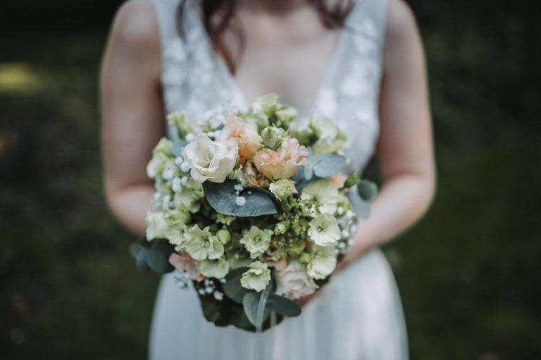 Die Braut, deren Kopf man nicht sieht, hält mit beiden Händen ihren Brautstrauß in die Kamera