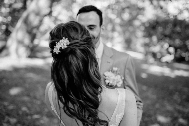 Schwarz-Weiß-Aufnahme eines Hochzeitspaares, bei der sich beide gegenüber stehen und anlächeln. Fotografiert von hinter der Braut.