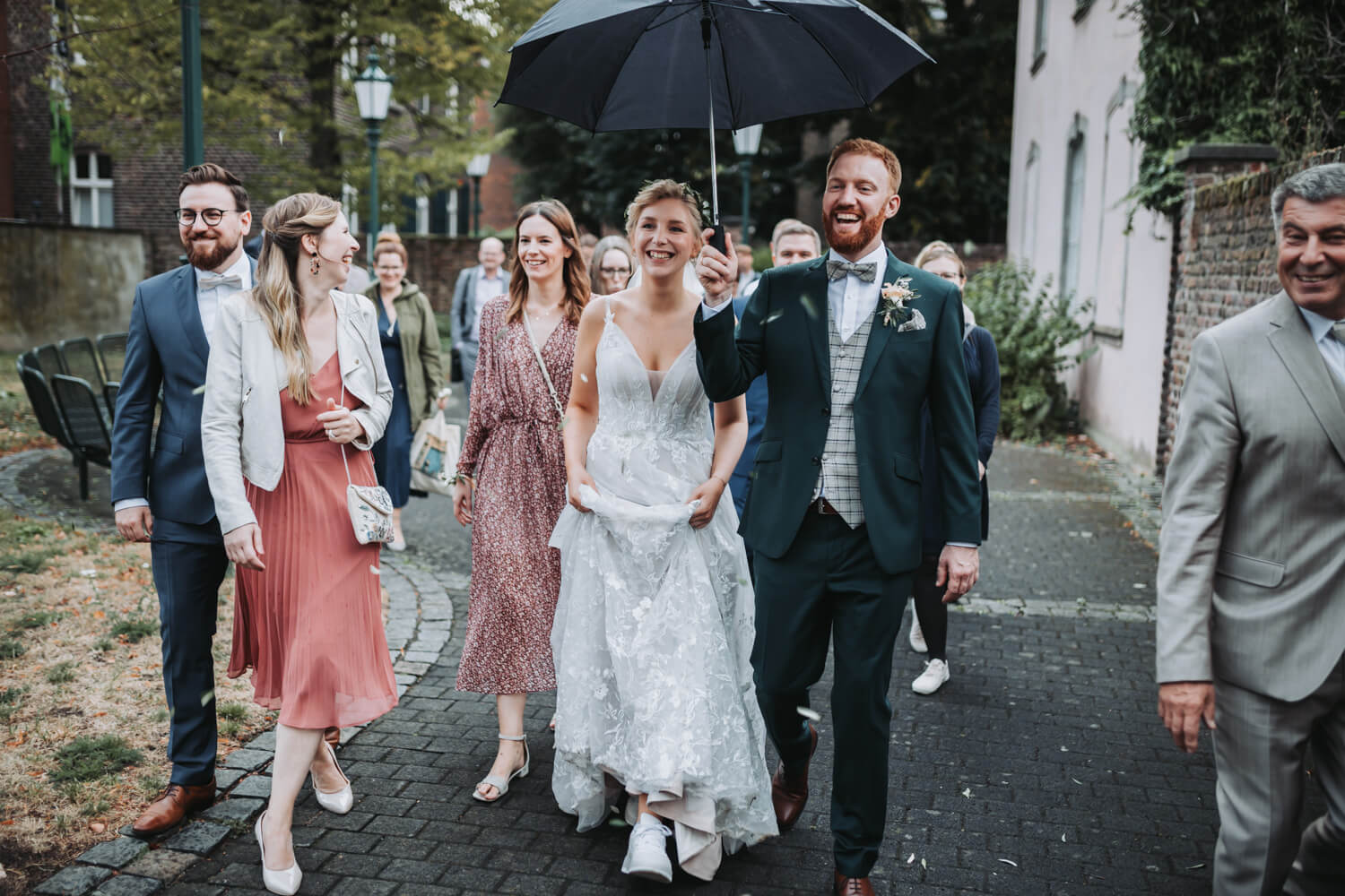 Hochzeitspaar geht mit einem Regenschirm und umgeben von ihren Gästen auf den Fotografen zu